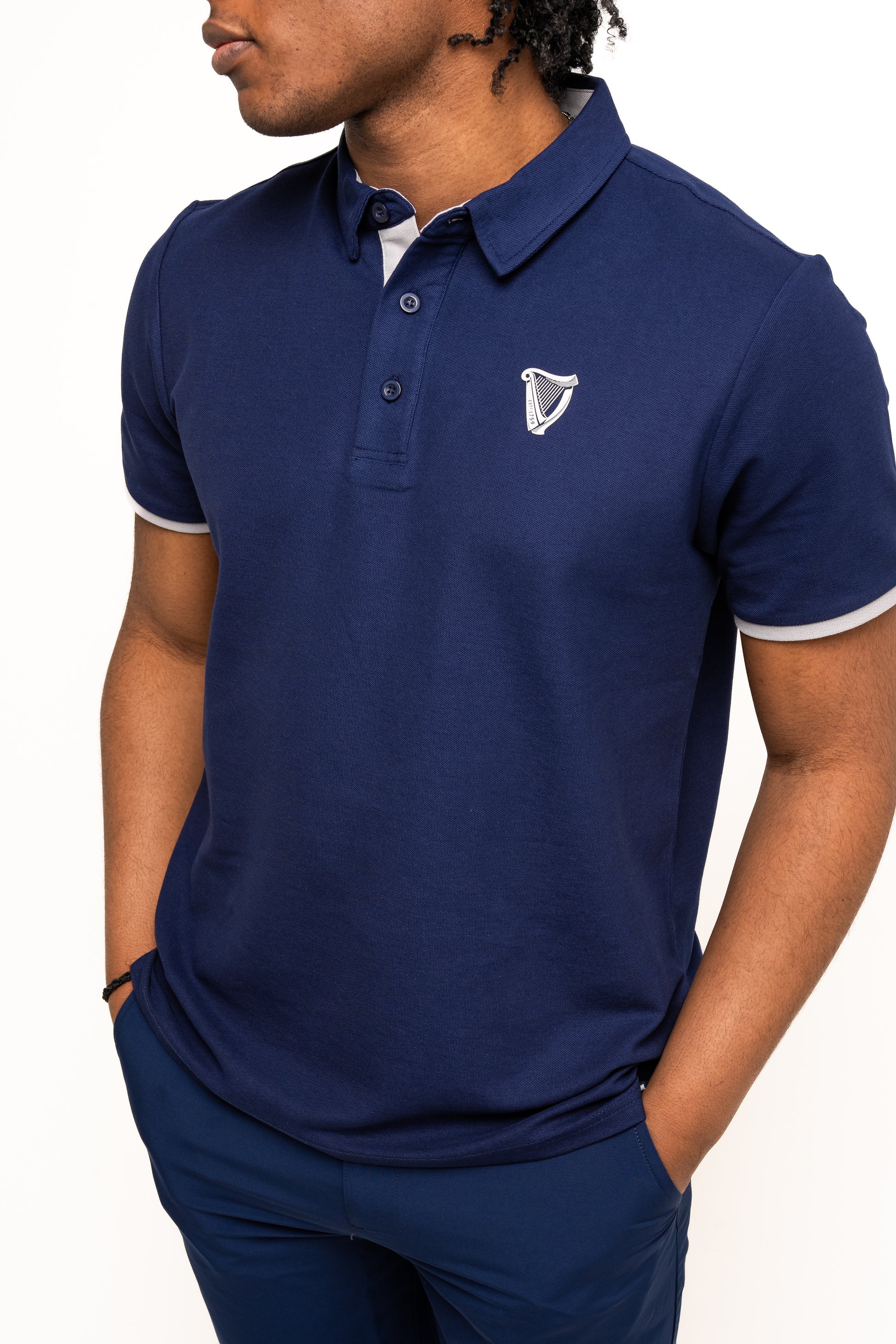 A person wears the Guinness Performance Polo - Navy by Guinness Webstore UK, made from premium 210gsm double pique fabric with a small white harp logo on the left chest, hands in pockets, shown from shoulders to waist.
