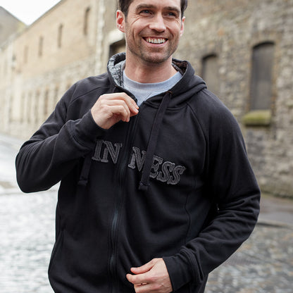 A person stands outdoors on a cobblestone street, smiling and wearing a dark Guinness Vintage Black & Grey Label Lined Hoodie from the Guinness Webstore UK, with the word "INNESS" visible on the front.