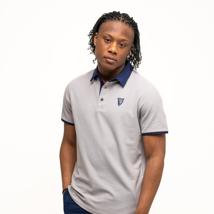 A person stands against a plain white background wearing the Guinness Performance Polo - Grey by Guinness Webstore UK, featuring a dark collar and sleeves, paired with navy blue shorts and one hand in their pocket.