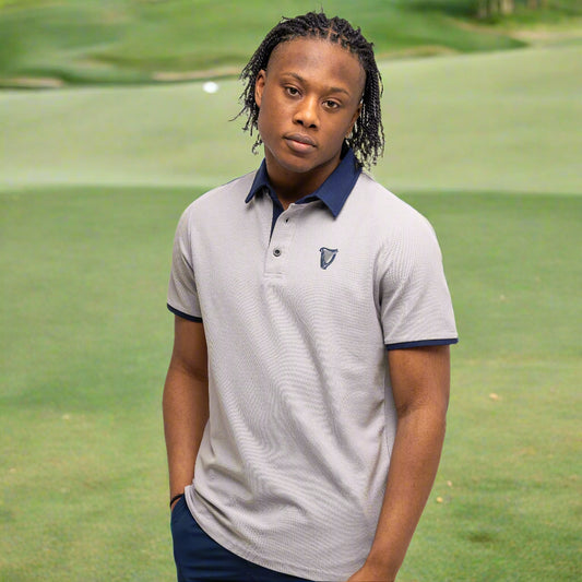 On a golf course, a person wears the Guinness Performance Polo - Grey from Guinness Webstore UK, made from premium 210gsm double pique fabric, paired with navy pants, and looks at the camera with a neutral expression.