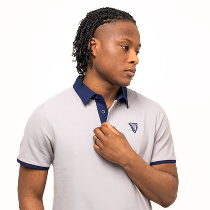 A person models the Guinness Performance Polo - Grey from Guinness Webstore UK, featuring a light grey body with dark blue collar and sleeve trim, made from premium double pique fabric, standing against a plain white background.