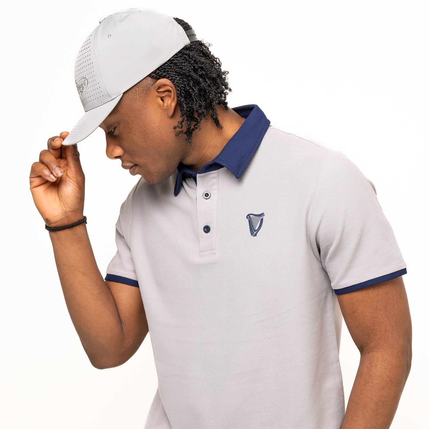 A man wearing the Guinness Performance Polo - Grey from Guinness Webstore UK with navy trim and a matching light grey cap, adjusting the brim of his hat as he looks down.