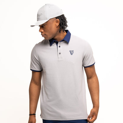 A man wears the Guinness Performance Polo - Grey from Guinness Webstore UK, featuring a dark collar and premium double pique fabric, paired with navy shorts and a gray cap, standing against a white background while looking left.