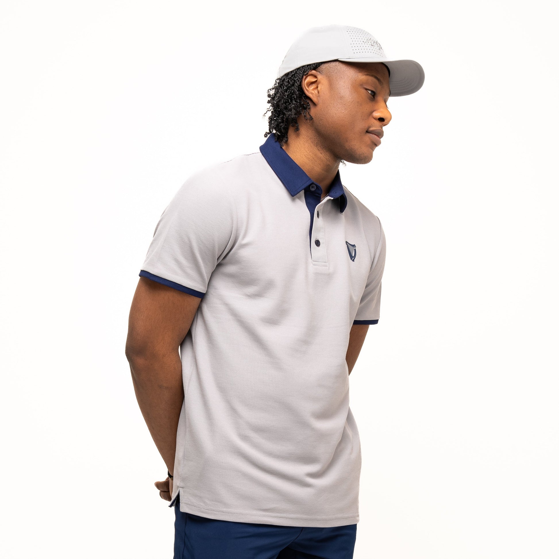 A man in the Guinness Webstore UK’s Guinness Performance Polo - Grey, paired with navy shorts and a gray cap, stands with his hands behind his back against a plain white background.