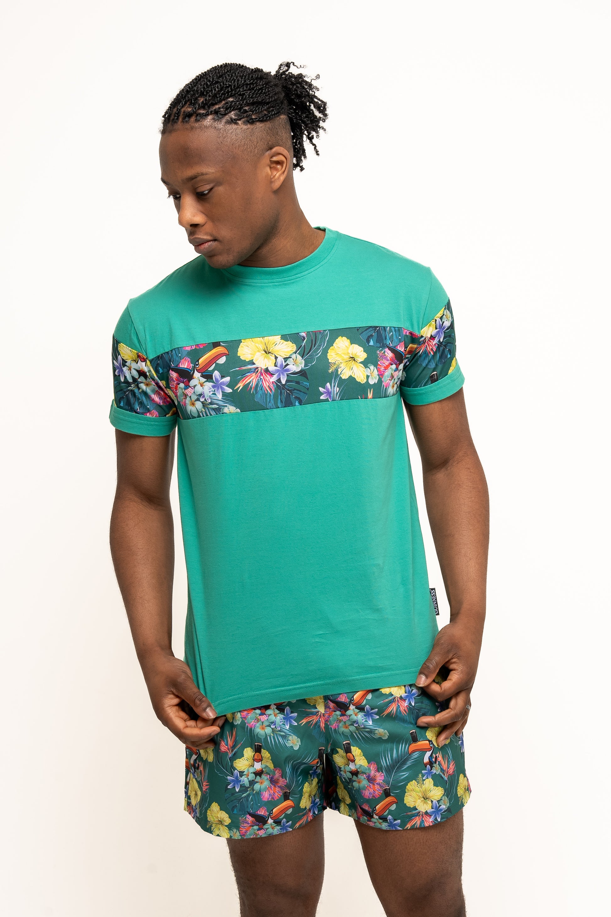 A man wearing a green t-shirt and Guinness Toucan Hawaiian Green Beach Shorts by Guinness UK stands against a plain white background, looking down to his left.