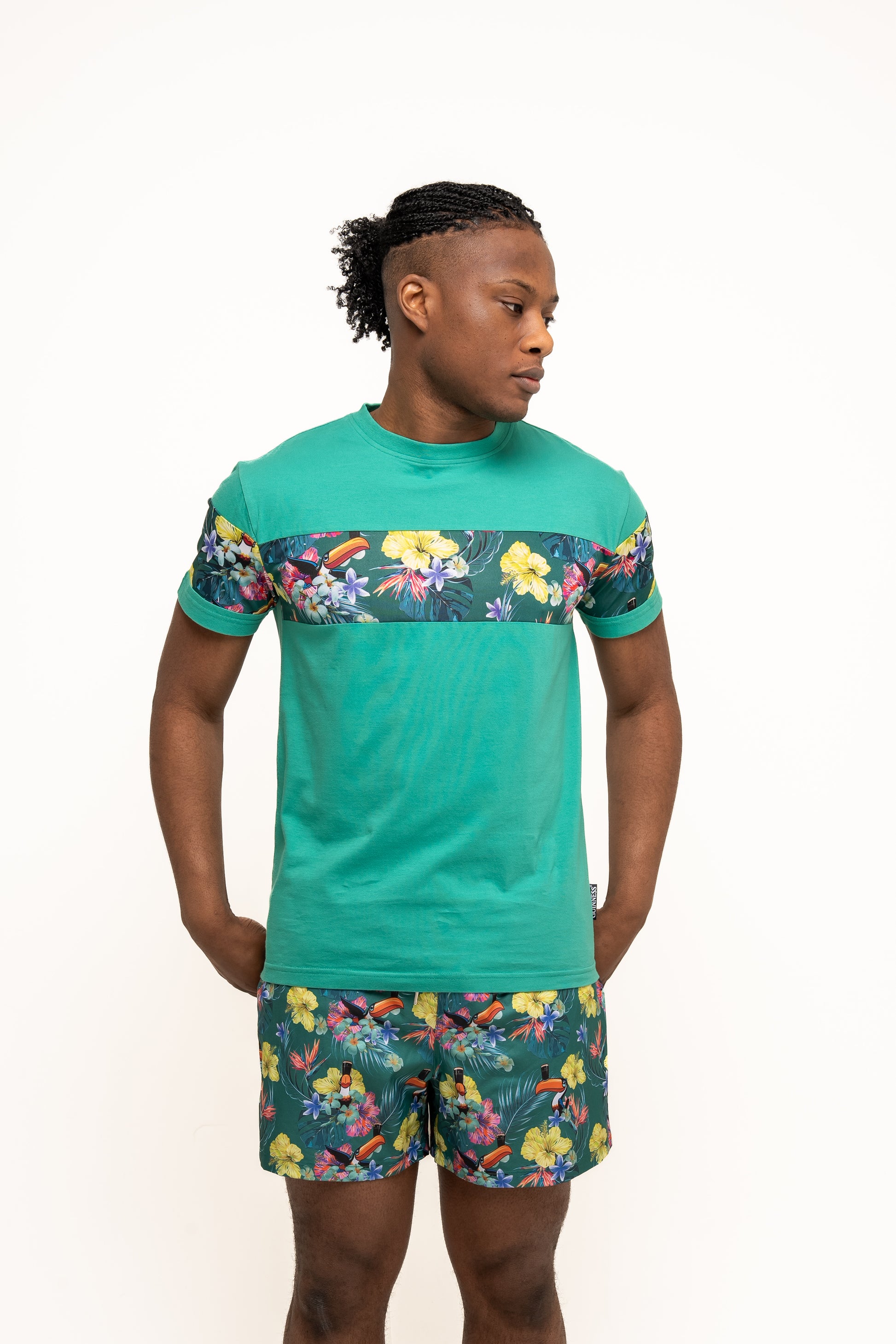 A person wears Guinness Toucan Hawaiian Green Beach Shorts by Guinness UK, a matching set with green t-shirt and shorts featuring floral prints and the iconic Guinness Toucan, looking to the side against a plain white background.
