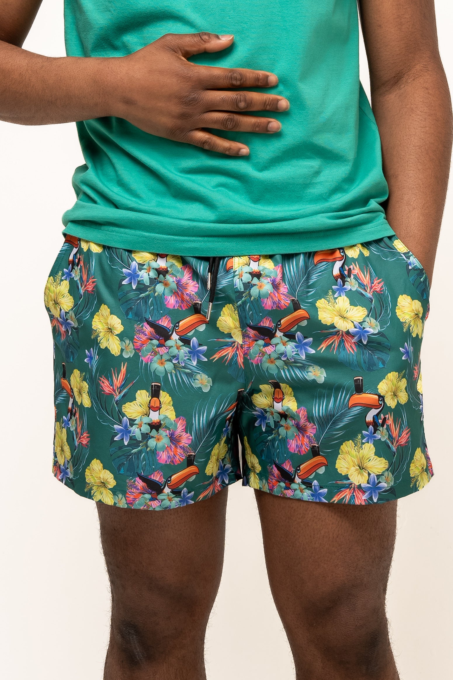 A person wearing Guinness UK Guinness Toucan Hawaiian Green Beach Shorts and a green shirt, with one hand on their stomach and the other in their pocket.