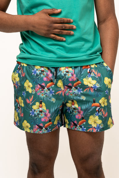 A person wearing Guinness UK Guinness Toucan Hawaiian Green Beach Shorts and a green shirt, with one hand on their stomach and the other in their pocket.