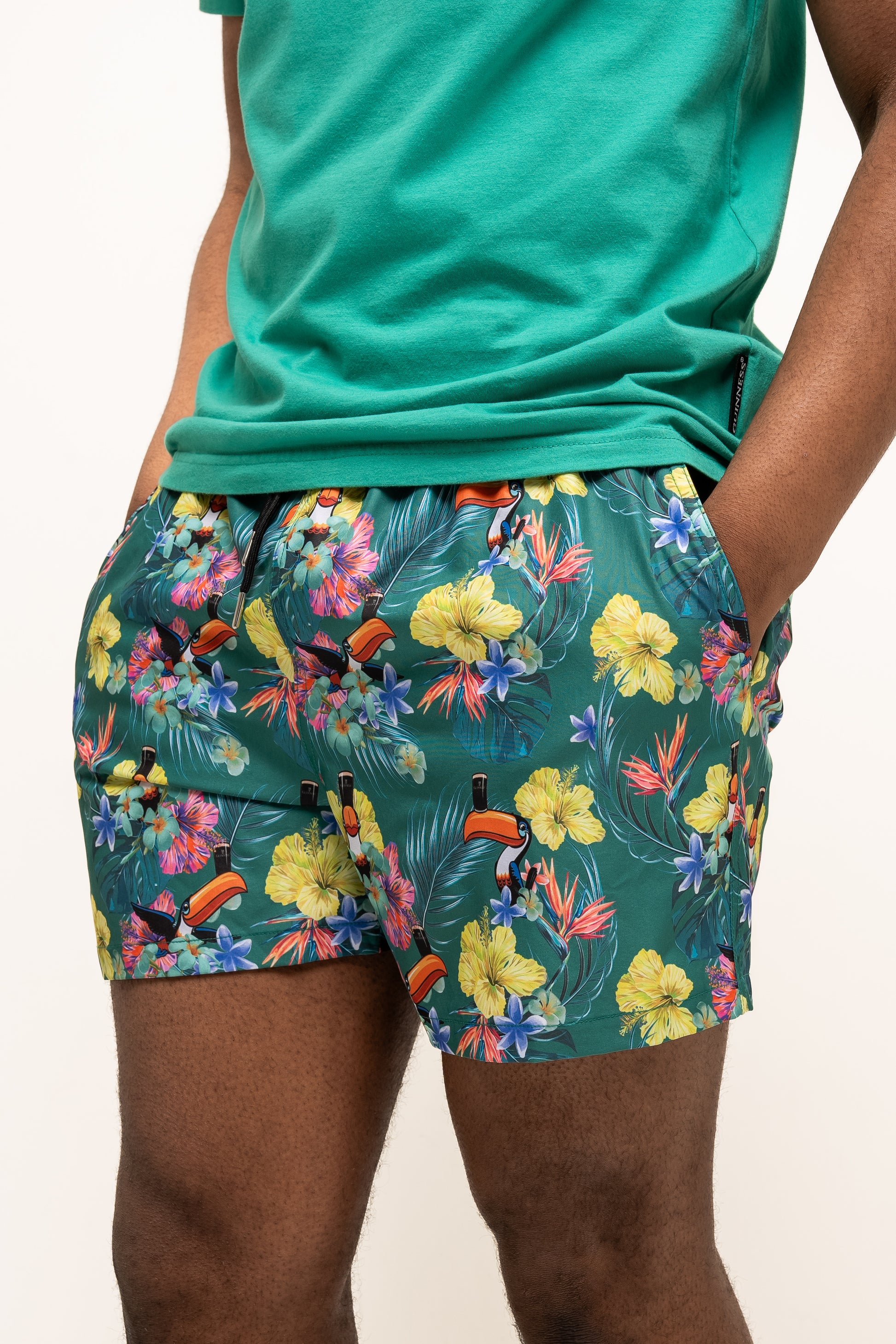 Wearing Guinness UK’s Guinness Toucan Hawaiian Green Beach Shorts and a matching tropical floral tee, someone stands with hands in pockets.