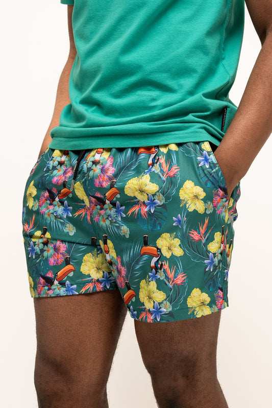 Wearing Guinness UK’s Guinness Toucan Hawaiian Green Beach Shorts and a matching tropical floral tee, someone stands with hands in pockets.