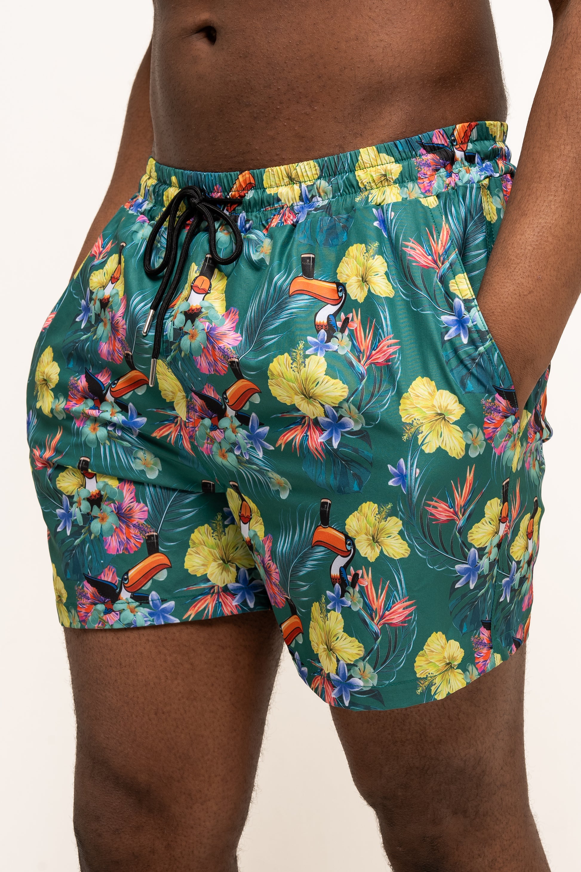 Someone pictured from mid-torso to mid-thigh wears Guinness UK’s Guinness Toucan Hawaiian Green Beach Shorts, featuring an eco-friendly, colorful tropical floral and toucan print.