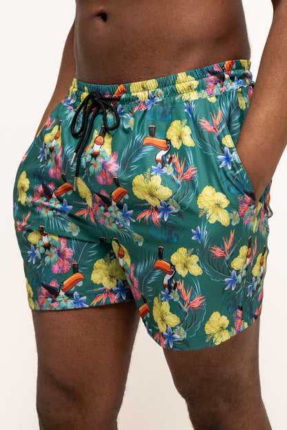 Someone pictured from mid-torso to mid-thigh wears Guinness UK’s Guinness Toucan Hawaiian Green Beach Shorts, featuring an eco-friendly, colorful tropical floral and toucan print.