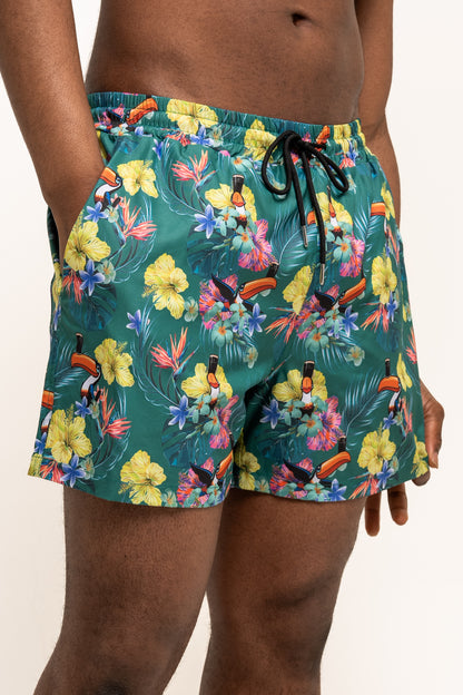 A person wearing Guinness Toucan Hawaiian Green Beach Shorts by Guinness UK, featuring a tropical floral and toucan print, standing with hands in pockets.