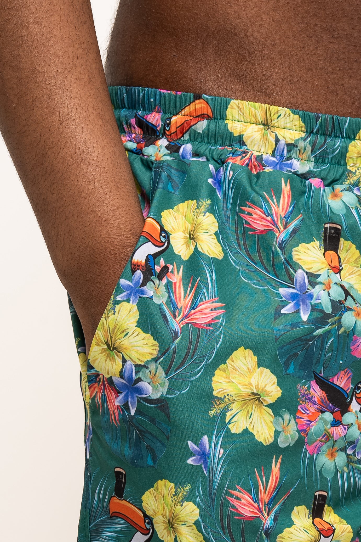 Close-up of a hand in the pocket of Guinness UK’s Guinness Toucan Hawaiian Green Beach Shorts, featuring a colorful tropical floral and toucan print.