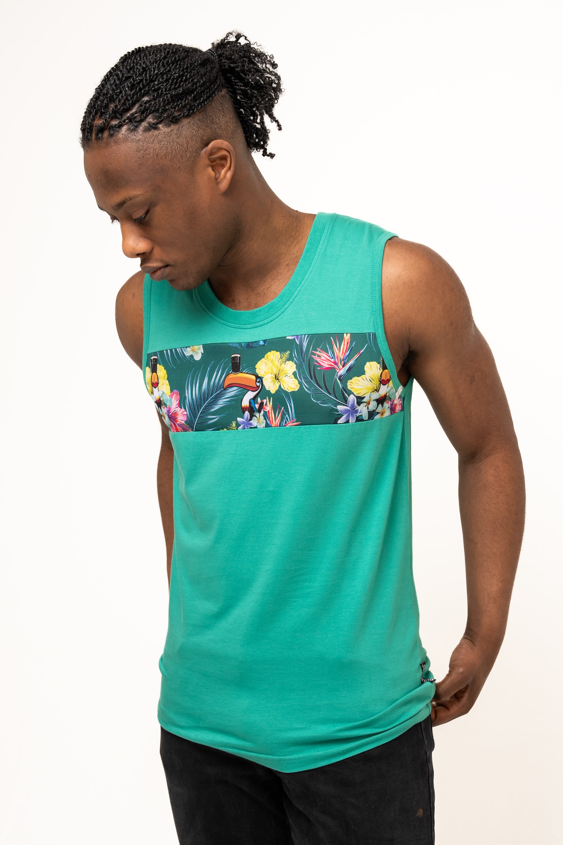 A man wears the Guinness Toucan Hawaiian Green Tank from Guinness Webstore UK, featuring a tropical floral and toucan print across the chest, standing against a plain light background and looking down with laid-back, paradise vibes.