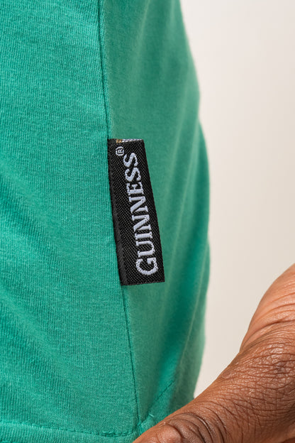 A close-up of a black and white "GUINNESS" label sewn onto the side seam of the Guinness Toucan Hawaiian Green Tank by Guinness Webstore UK, next to a person's hand.