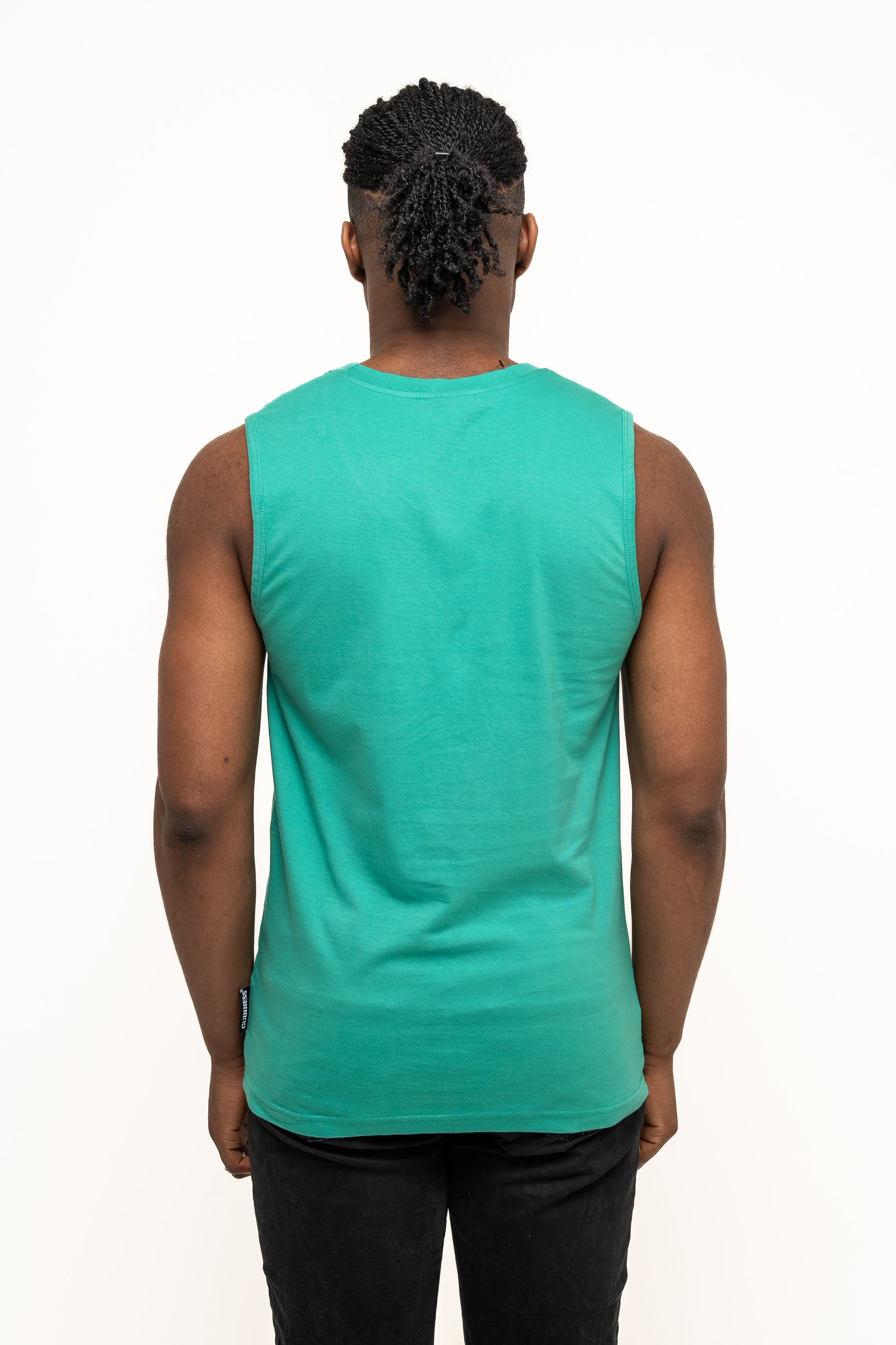 A person with braided hair stands with their back to the camera, wearing the Guinness Toucan Hawaiian Green Tank by Guinness Webstore UK and black pants against a plain white background.