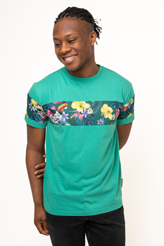 A person wearing the Guinness Toucan Hawaiian Green T-Shirt by Guinness UK, featuring a floral and toucan print stripe across the chest and sleeves, stands smiling against a plain white background.