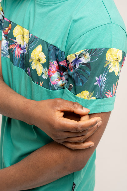 A person wearing the Guinness UK Guinness Toucan Hawaiian Green T-Shirt, featuring colorful tropical flora on the sleeve and chest, is adjusting the sleeve with one hand.