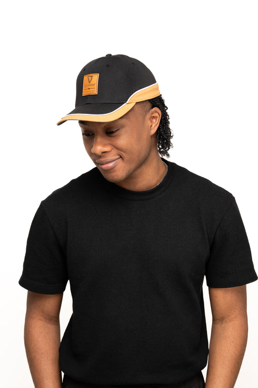 A person in a black t-shirt and the Guinness Black Cap with Caramel Edge from Guinness Webstore UK, featuring a tan leatherette patch, looks down and smiles against a white background.