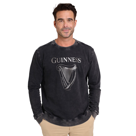 A man faces forward wearing the Guinness Metallic Print Trademark Sweatshirt by Guinness Webstore UK, featuring a metallic harp graphic and sustainable cotton, paired with beige pants against a plain white background.