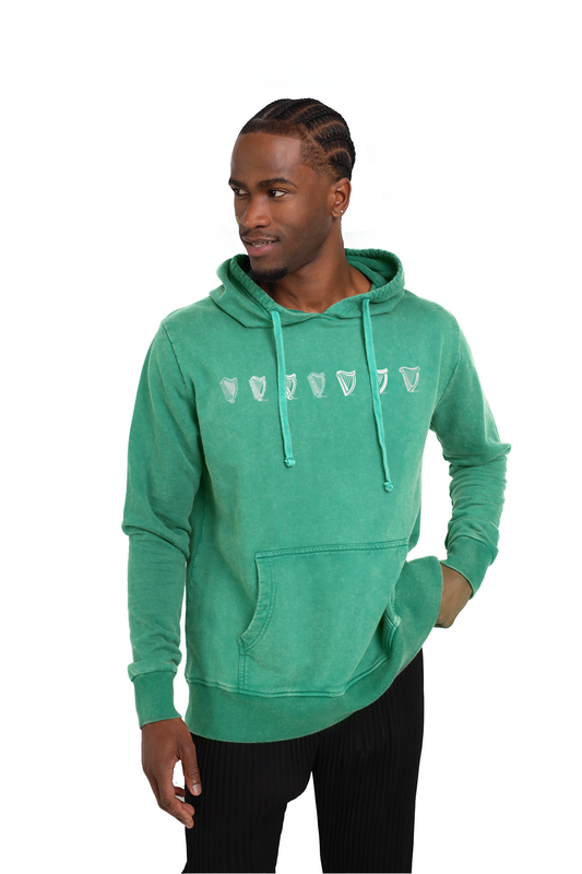 A person in a green Guinness Evolution Harp Hoodie, adorned with geometric patterns and the subtle Guinness Harp logo, stands against a white background, gazing thoughtfully to the side with hands in pockets.