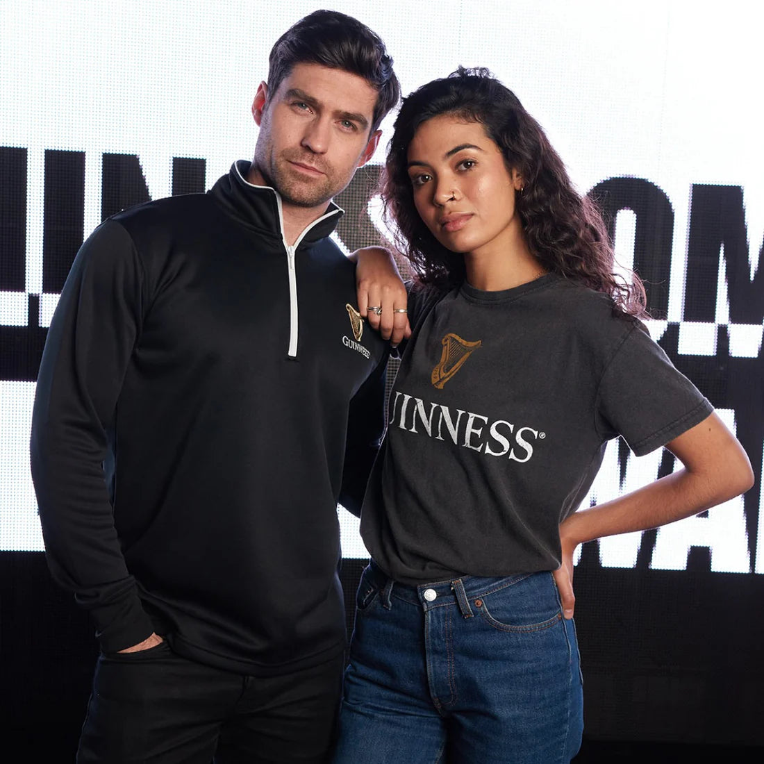 A man and woman pose with serious expressions while wearing Guinness UK-branded clothing; the man sports a black Guinness Harp Half Zip Top, showcasing technical performance fabric, and the woman wears a grey t-shirt.