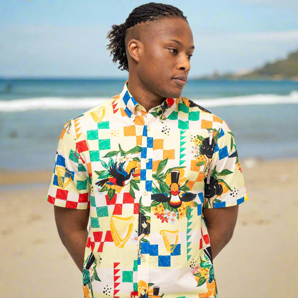 On the beach, a person enjoys the comfort of breathable woven cotton in a colorful Guinness Toucan Beach Shirt from Guinness Webstore UK, with the ocean and shoreline in the background.
