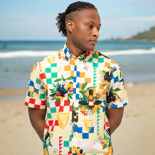 On the beach, a person enjoys the comfort of breathable woven cotton in a colorful Guinness Toucan Beach Shirt from Guinness Webstore UK, with the ocean and shoreline in the background.