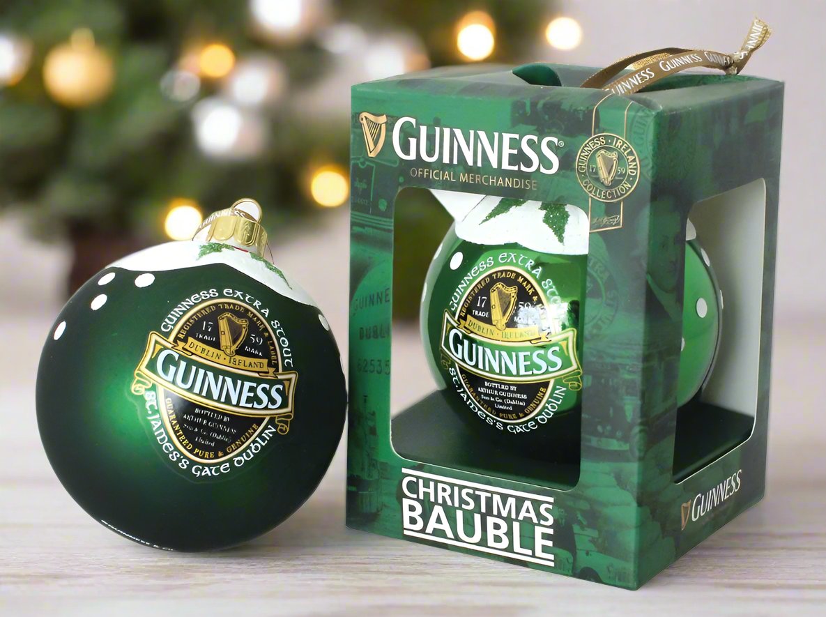 The Guinness Ireland Christmas Bauble, from the renowned brand Guinness, showcases the classic logo on a green bauble. Presented both inside and outside its packaging, it is an ideal gift for beer enthusiasts, set against the gentle blur of a festive tree backdrop.