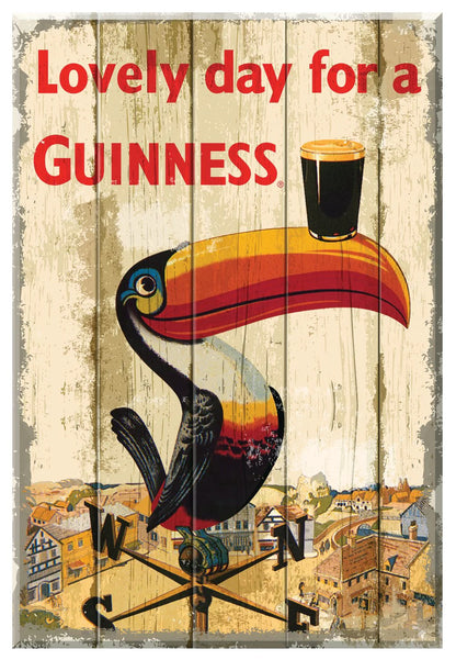 The Guinness UK Nostalgic Sign - Toucan Weathervane features a vintage advertisement with a toucan balancing a pint of Guinness on its beak and the text "Lovely day for a Guinness" against a wood background. This charming piece is part of the nostalgic collection, making it an ideal wall art addition.