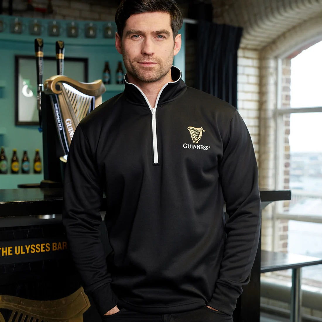 In a bar adorned with shelves of bottles and a harp decoration, a man stands wearing the Guinness UK Guinness Harp Half Zip Top in black, its technical performance fabric ensuring his comfort.