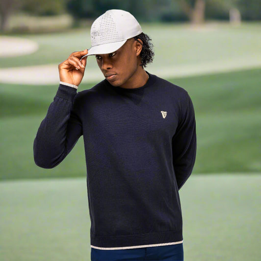 A man in a Guinness Performance Cotton Sweater - Navy from Guinness Webstore UK and a light cap stands on a golf course, holding the brim of his cap with one hand.