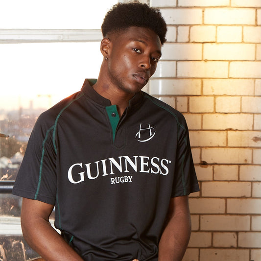 A man wearing a Guinness Black & Green Short Sleeve Rugby Jersey sits indoors by a window and white tiled wall, illuminated by natural light.
