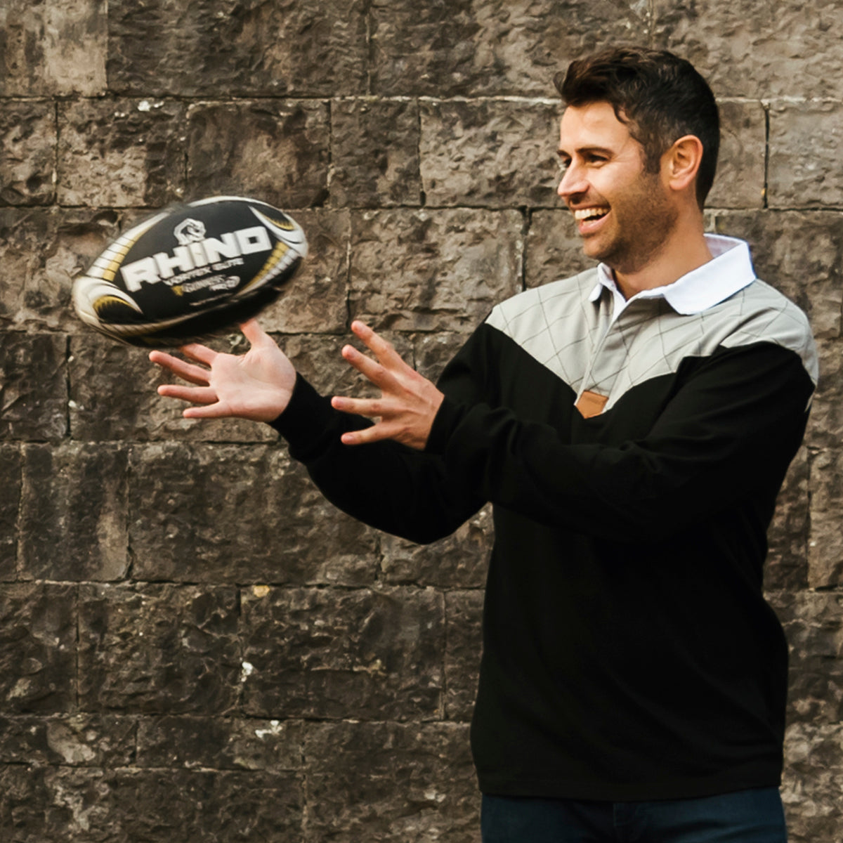 In front of a stone wall, a man smiles as he catches a rugby ball, wearing the Guinness Heritage Long Sleeve Rugby Jersey in black and gray, made from 100% cotton.