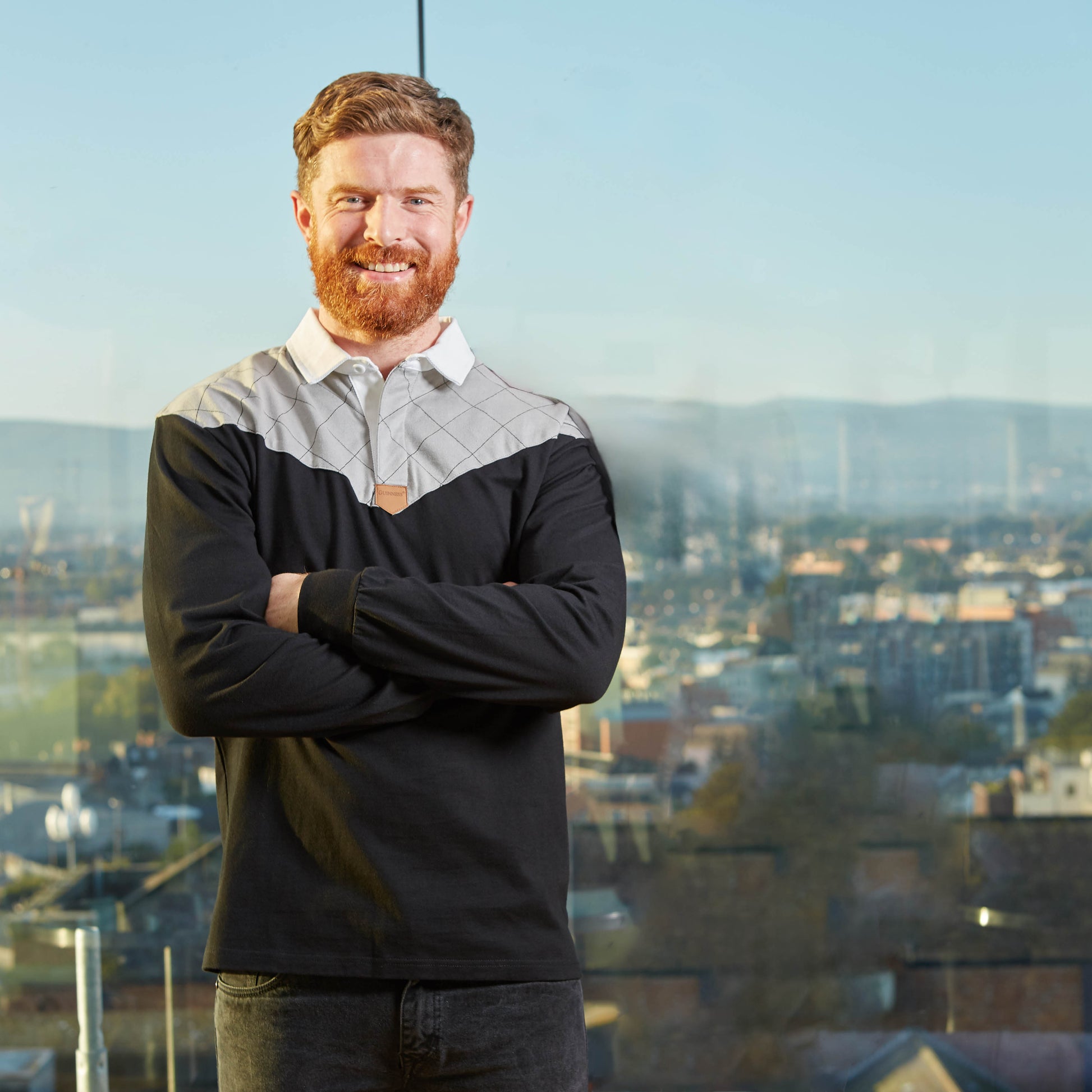 A red-haired, bearded man stands with arms crossed in a black and grey Guinness Heritage Long Sleeve Rugby Jersey, made from 100% cotton, in front of a large window with a cityscape view.