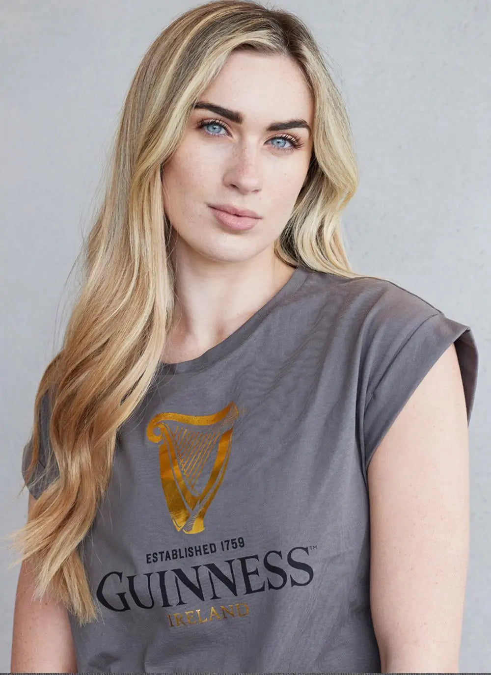 Woman with long blonde hair wearing a gray Guinness Webstore UK Gold Foil Harp T-shirt, looking directly at the camera.