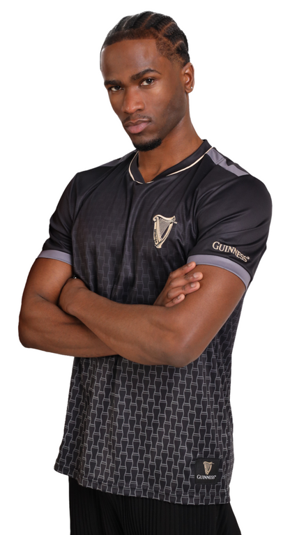 A man in a black and grey Guinness Football Jersey, featuring the iconic Guinness badge, stands with arms crossed, looking directly at the camera.