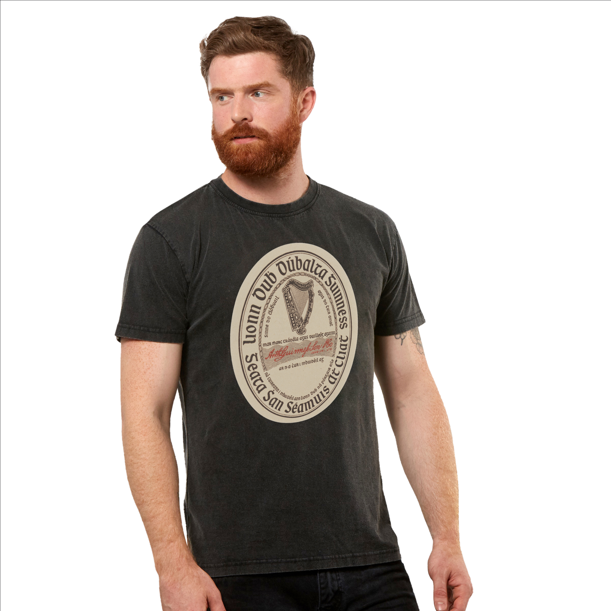 A man with a beard and short hair wears a black Guinness Gaelic Label Black Premium T-Shirt from Guinness UK, made of 100% cotton. The large circular design in the center features text and a graphic. He is looking to his left, showcasing the unisex style.