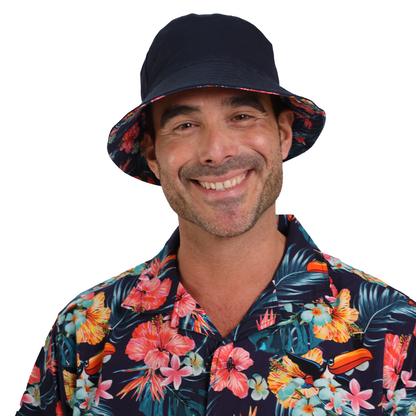 A person smiling, wearing a Guinness Hawaiian Reversible Bucket Hat from the Guinness Webstore UK and a floral patterned shirt with a dark background.