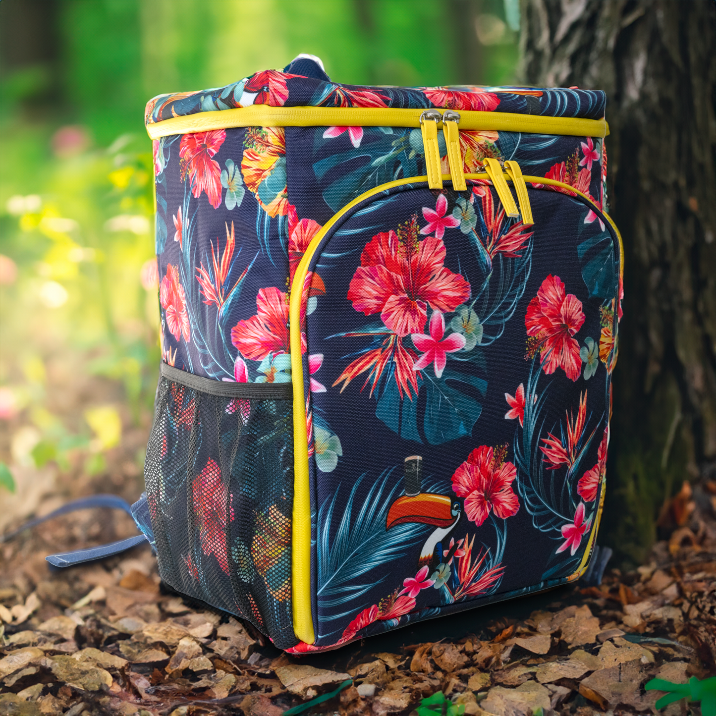 A backpack adorned with a floral pattern, featuring yellow zippers and mesh side pockets, rests on the forest floor, mirroring the charm of a Guinness Toucan Hawaiian Cooler Bag from the Guinness Webstore UK amid the fallen leaves and trees.