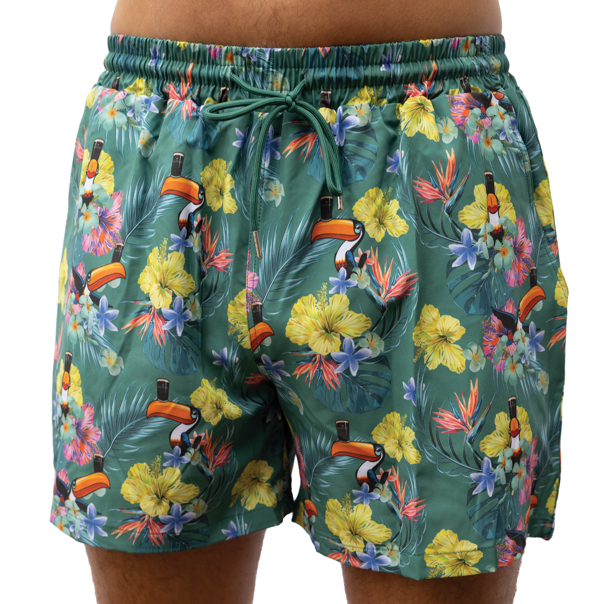 A person wearing Guinness Toucan Hawaiian Green Beach Shorts by Guinness UK, showcasing a vibrant tropical pattern with toucans and various flowers.