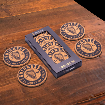 A set of four Guinness Harp cork coasters rests on a wooden table. Each coaster features the iconic logo, and they originated from Guinness, as indicated by a nearby box labeled "Guinness Harp Cork Coaster Set - 4 Pack.