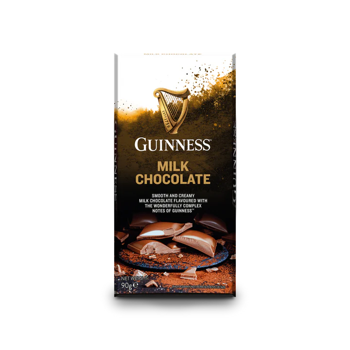 A Guinness Luxury Milk Chocolate Bar with chunks of milk chocolate displayed on a brown backdrop. The packaging, which is part of the official Guinness merchandise, showcases the iconic logo and text that emphasizes the bar's rich flavor notes.