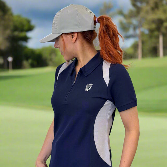 A woman in a blue Ladies Performance Polo from Guinness Webstore UK and a light gray cap stands on a golf course, looking to the side, with trees and green grass in the background.
