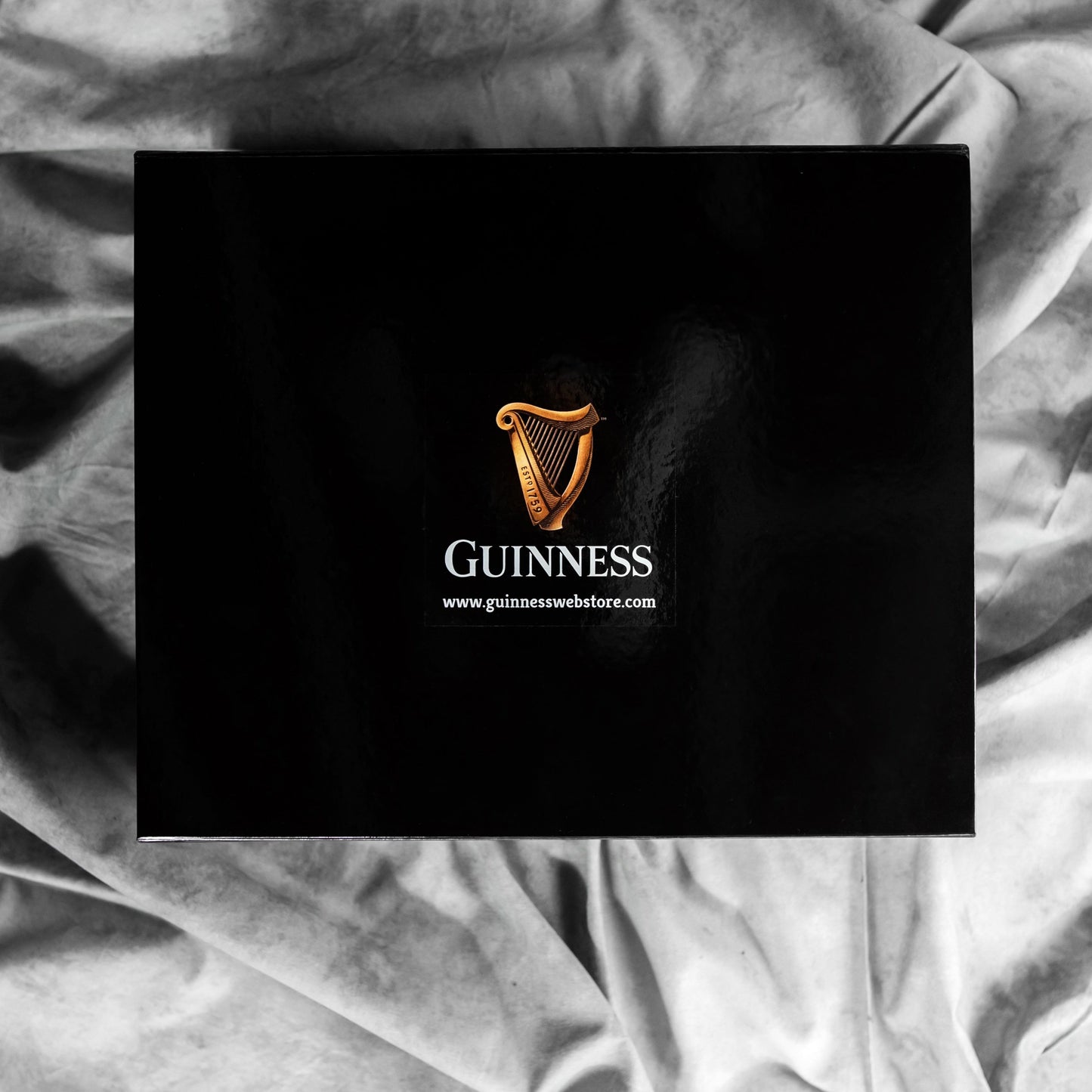 A Guinness Gift Box - Large, black in color and adorned with the iconic Guinness logo featuring a golden harp, is displayed against a backdrop of wrinkled gray fabric. Below the logo, the website "www.guinnesswebstore.com" is printed, suggesting that this could be a collector's edition from Guinness.