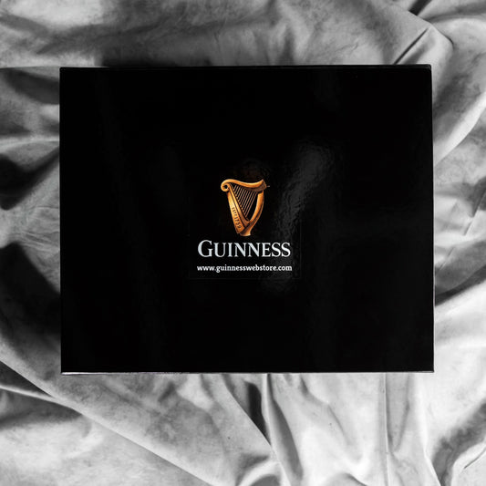 A Guinness Gift Box - Large, black in color and adorned with the iconic Guinness logo featuring a golden harp, is displayed against a backdrop of wrinkled gray fabric. Below the logo, the website "www.guinnesswebstore.com" is printed, suggesting that this could be a collector's edition from Guinness.