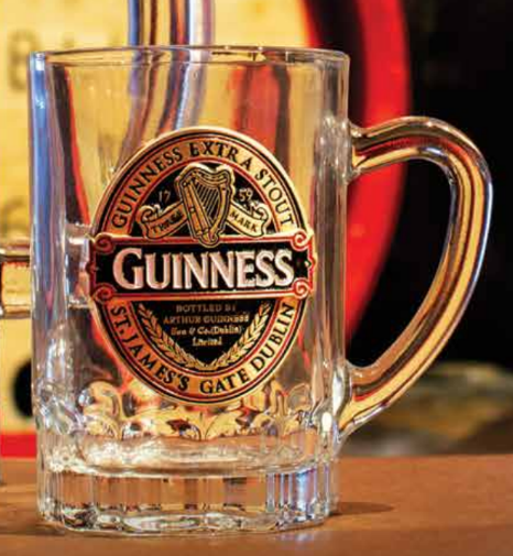 A clear glass mini tankard from the Guinness Classic collection by Guinness Webstore UK, adorned with the iconic Guinness logo and text, stands proudly on a wooden surface, set against a faintly blurred background.