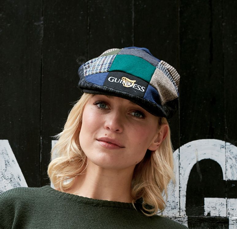 A woman wearing a colorful Guinness Tweed Cap with traditional Irish design, smiling slightly, against a black wooden background.