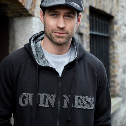 A man wearing a Guinness Vintage Black & Grey Label Lined Hoodie from the Guinness Webstore UK and a flat cap with the logo "Arthur Guinness" stands outside near a building with stone walls and barred windows.